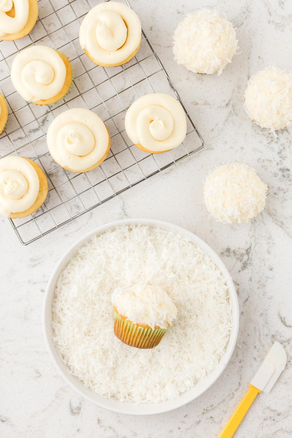 A cupcake is rolled in shredded coconut next to frosted cupcakes and coconut-covered cupcakes on a white countertop.