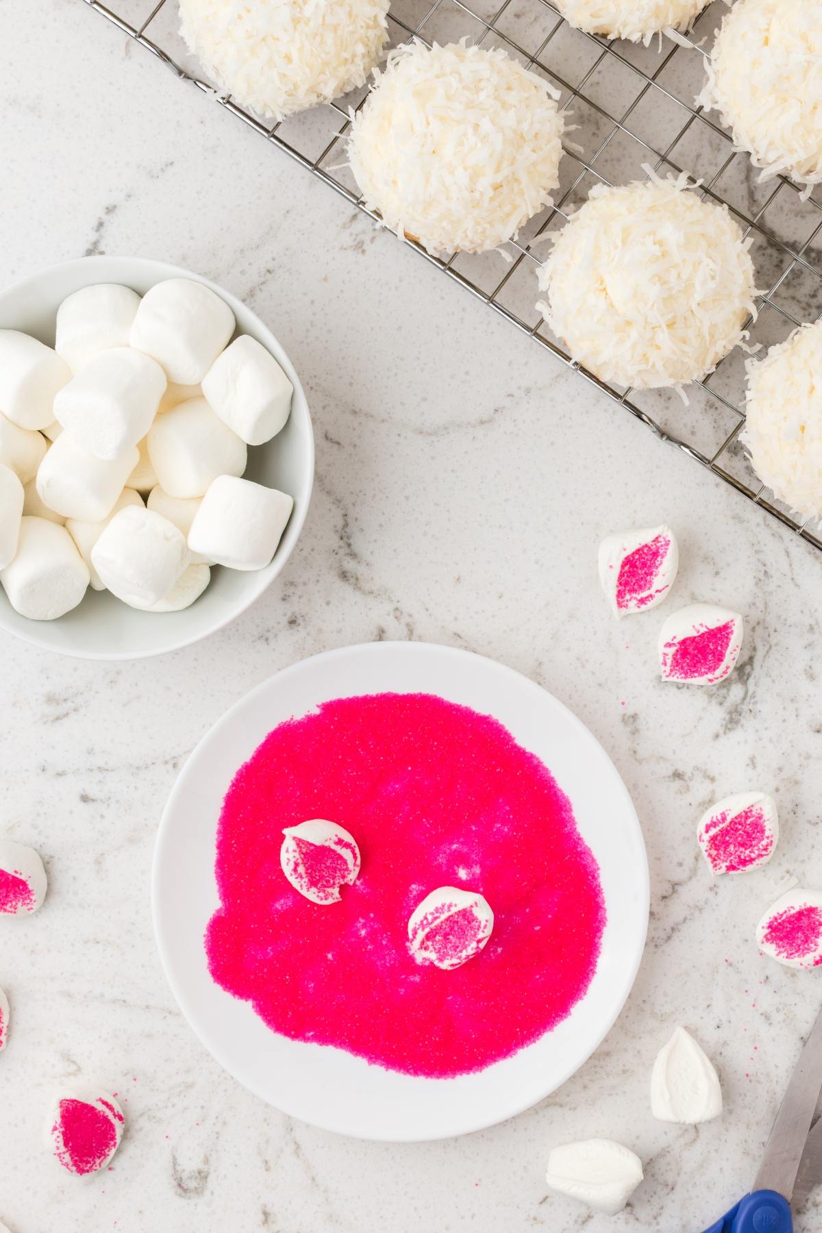 A plate with pink sugar and marshmallow halves, a bowl of marshmallows, and coconut treats on a cooling rack.