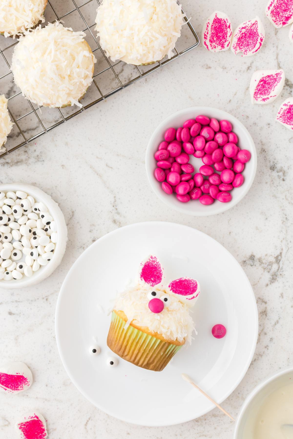 A bunny-themed cupcake with pink candy ears and nose sits on a plate, surrounded by candy and baking supplies.