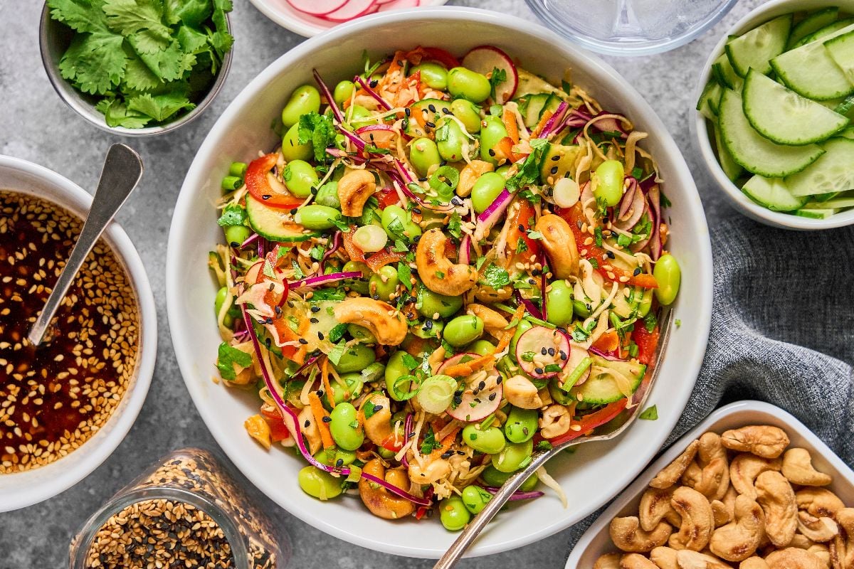 Colorful Cashew Crunch Edamame Salad with cashews, shredded veggies, and sesame seeds in a white bowl, surrounded by fresh toppings.