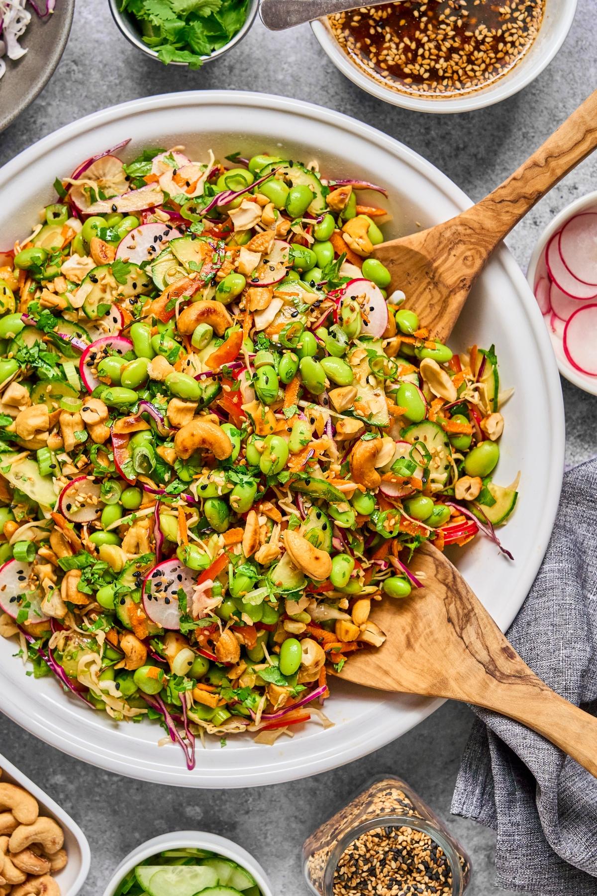 Colorful salad with edamame, cashews, radishes, and shredded veggies in a large bowl with wooden salad servers.