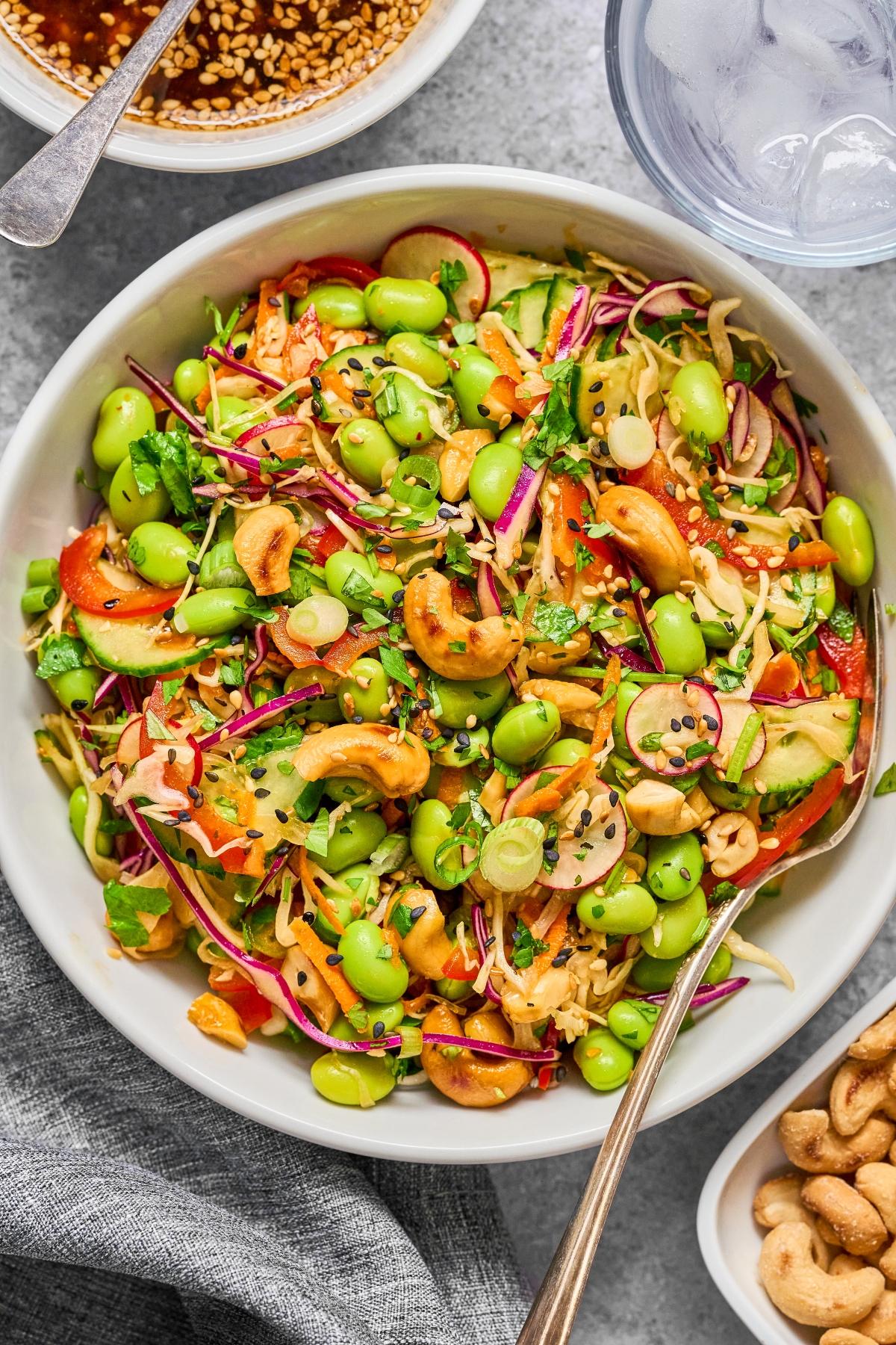 A colorful salad with edamame, cashews, shredded veggies, and black sesame seeds in a white bowl.