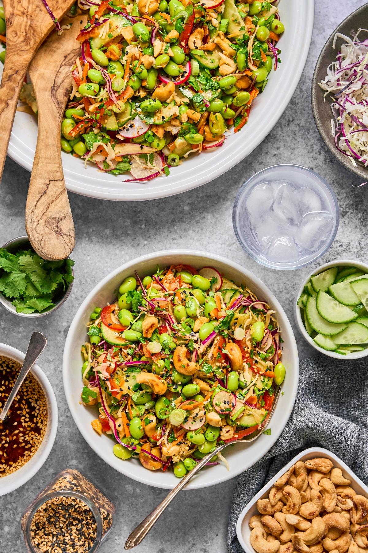 Colorful edamame salad with veggies and cashews, surrounded by dressing, cucumber, herbs, and a glass of ice water.