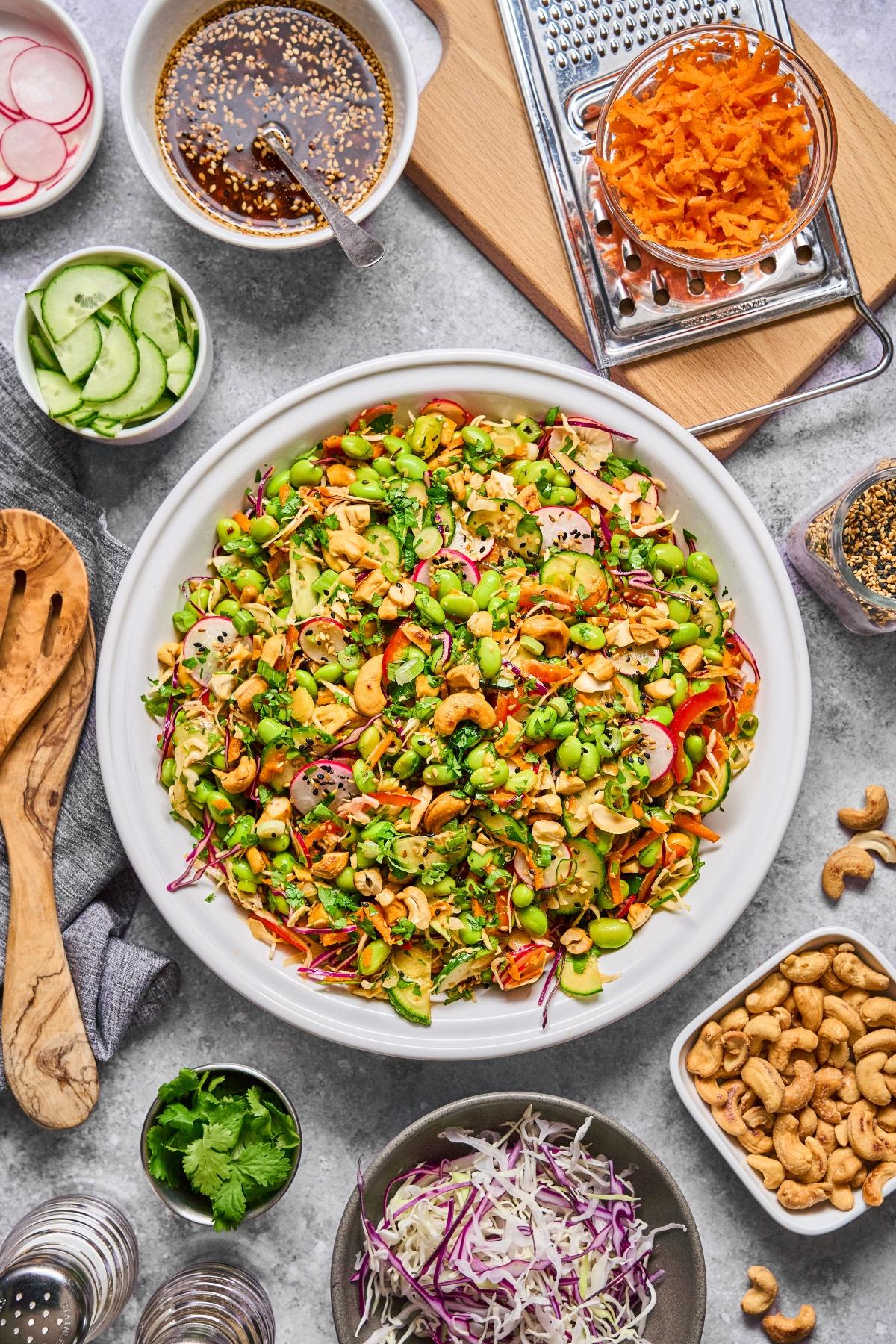 A colorful salad with mixed vegetables, cashews, and herbs, surrounded by bowls of toppings and dressing.