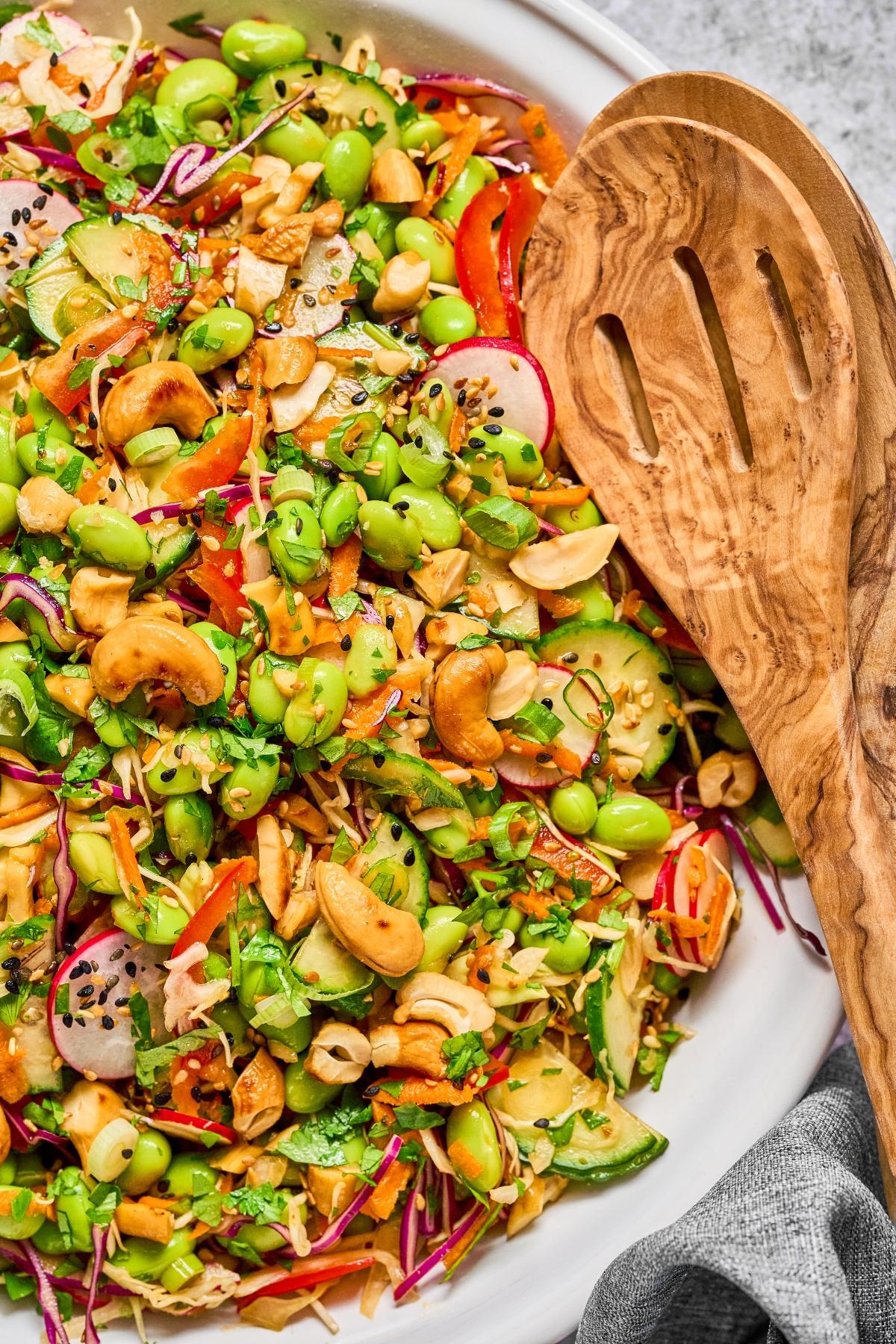 Colorful salad with edamame, cashews, sliced radishes, cucumbers, and veggies in a bowl with a wooden serving spoon.