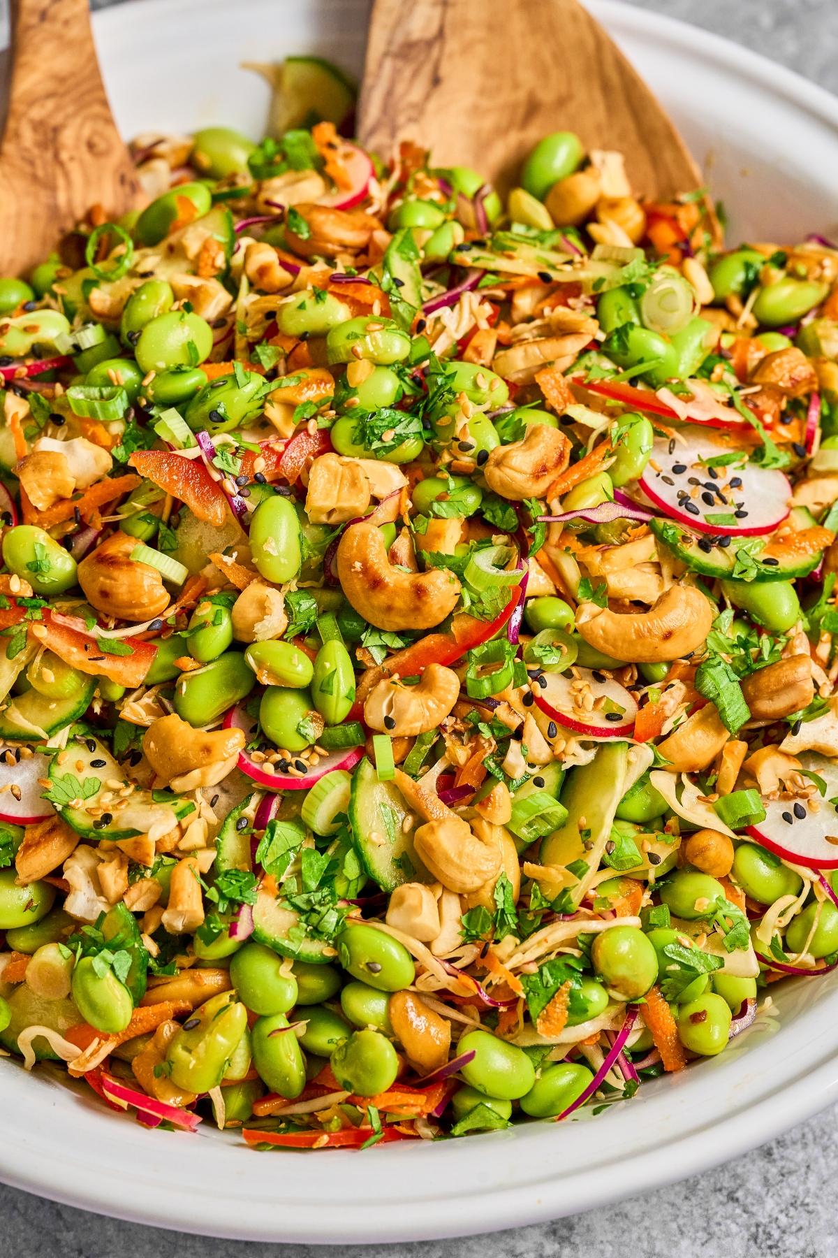 A colorful salad with edamame, radishes, cashews, carrots, herbs, and sesame seeds in a white bowl.
