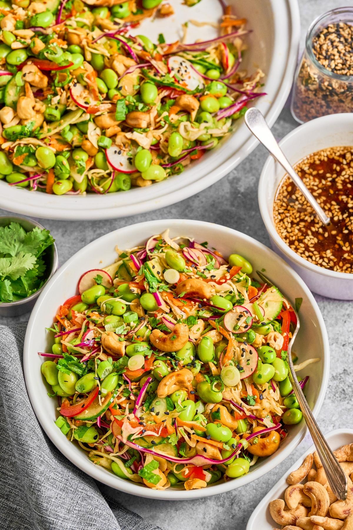 A bowl of colorful Asian salad with edamame, carrots, cabbage, cashews, and sesame seeds, with dressing on the side.