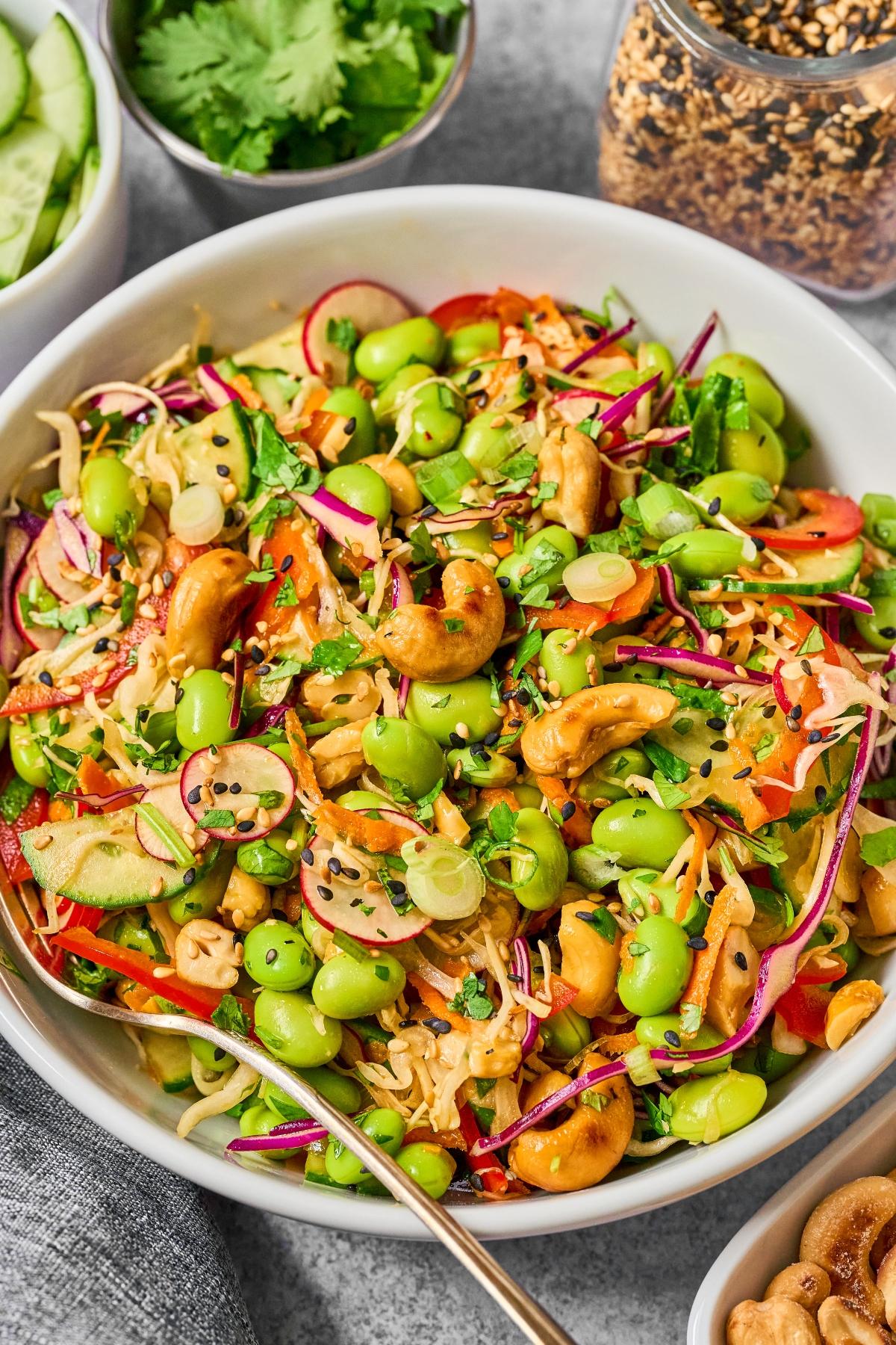 Colorful salad with edamame, cashews, sliced veggies, and sesame seeds in a white bowl with a gold fork.