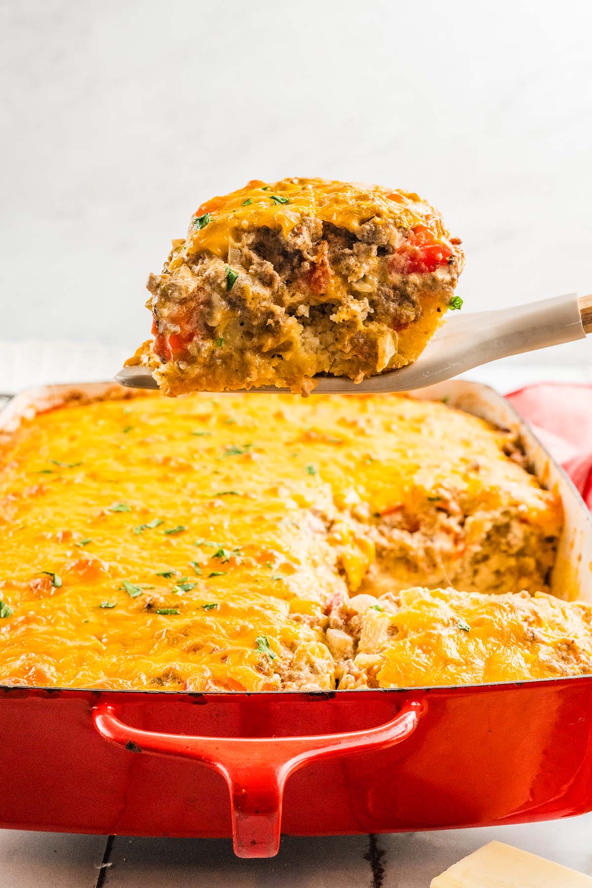 A serving of cheesy Cattle Drive Casserole is being lifted from a red baking dish filled with the savory, baked casserole.