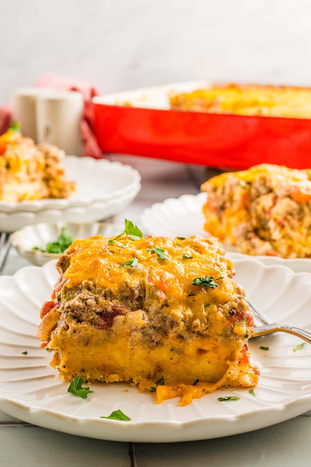 A slice of cheesy Cattle Drive Casserole on a white plate, garnished with parsley, with more hearty casserole in the background.
