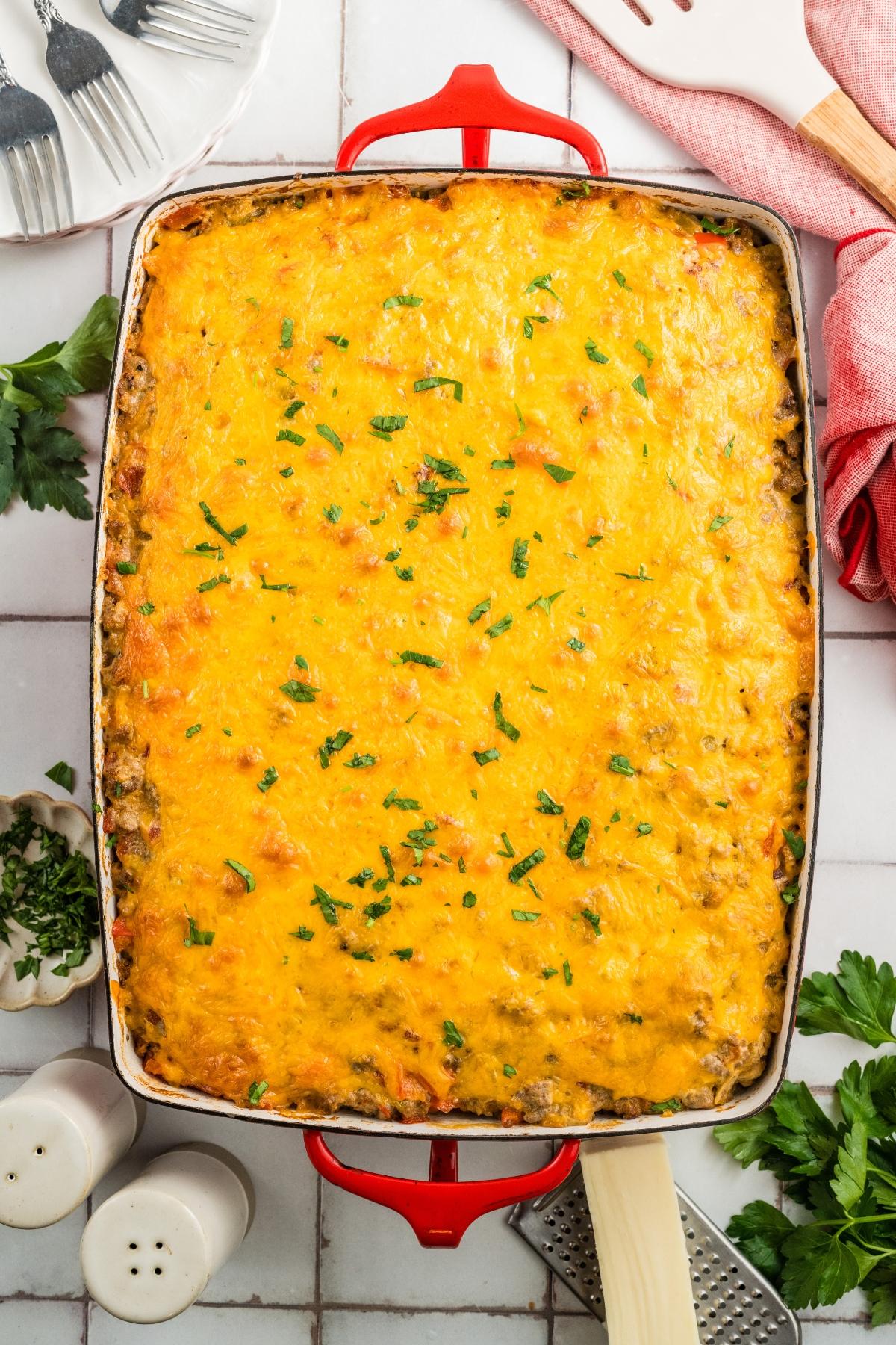A baked casserole topped with melted cheese, garnished with chopped herbs in a red baking dish.