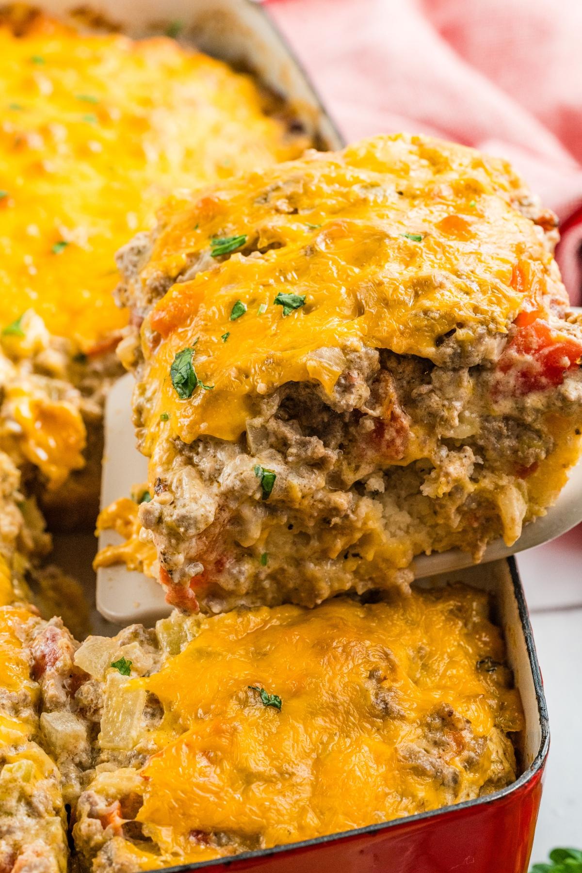 A serving of cheesy casserole with ground beef and vegetables being lifted from a baking dish.
