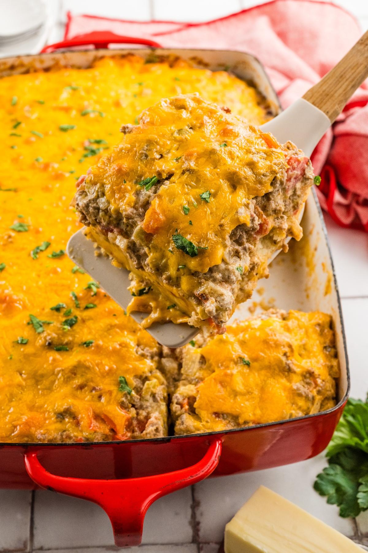 A spatula lifts a cheesy baked casserole from a red dish, topped with melted cheddar and herbs.