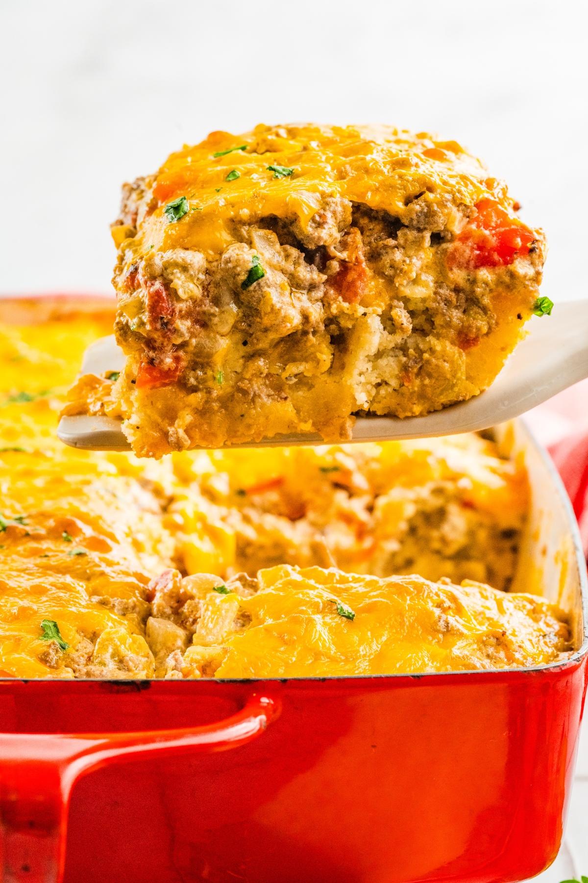 A spatula lifts a cheesy baked casserole from a red dish, showing meat and vegetables inside.