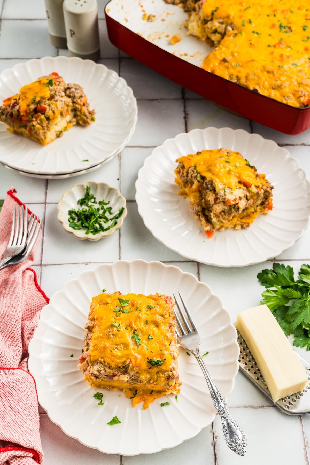 Three plates with cheesy casserole squares, garnished with parsley, beside a baking dish and cheese grater.