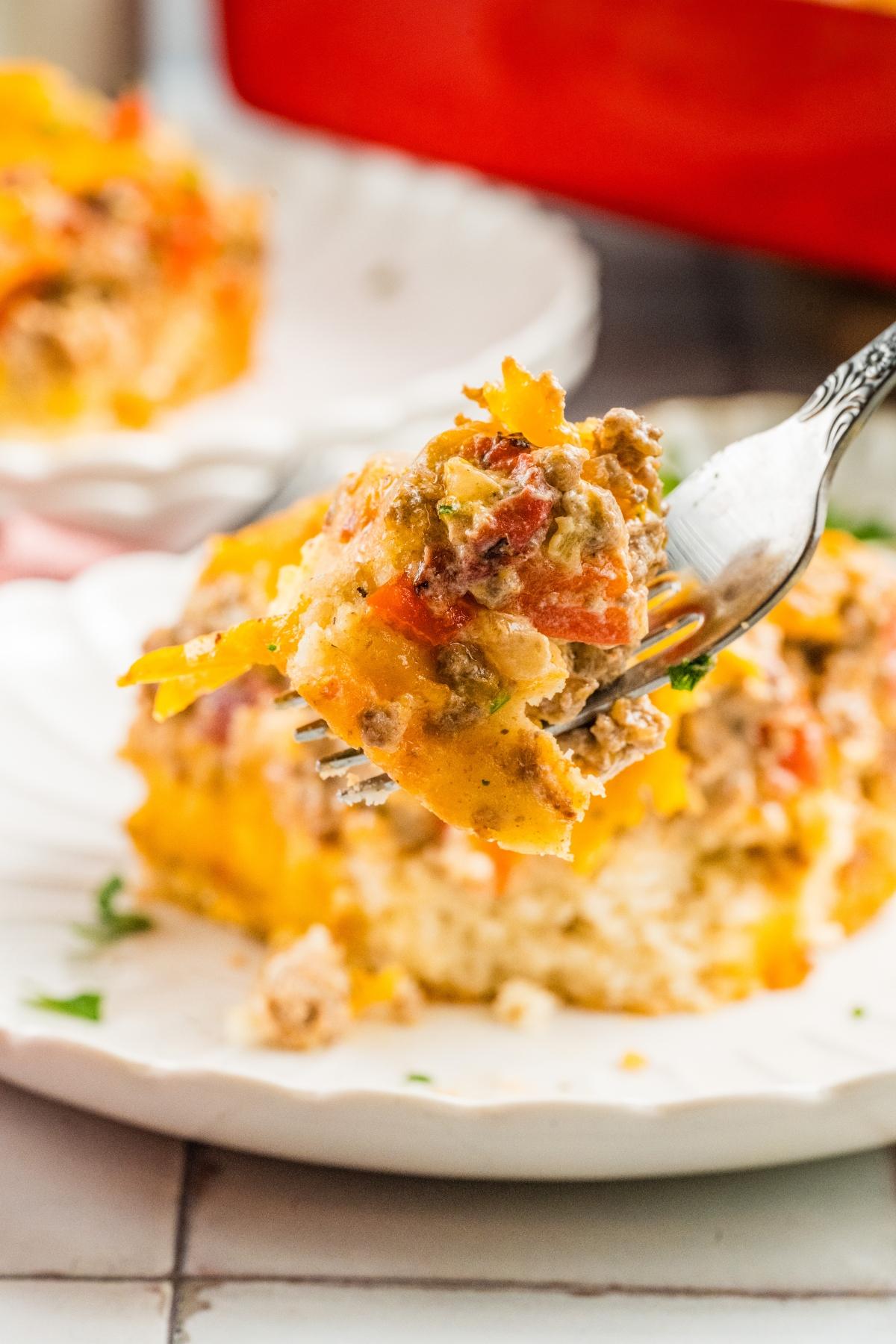 A fork holds a bite of cheesy casserole with ground meat and vegetables above a serving on a white plate.