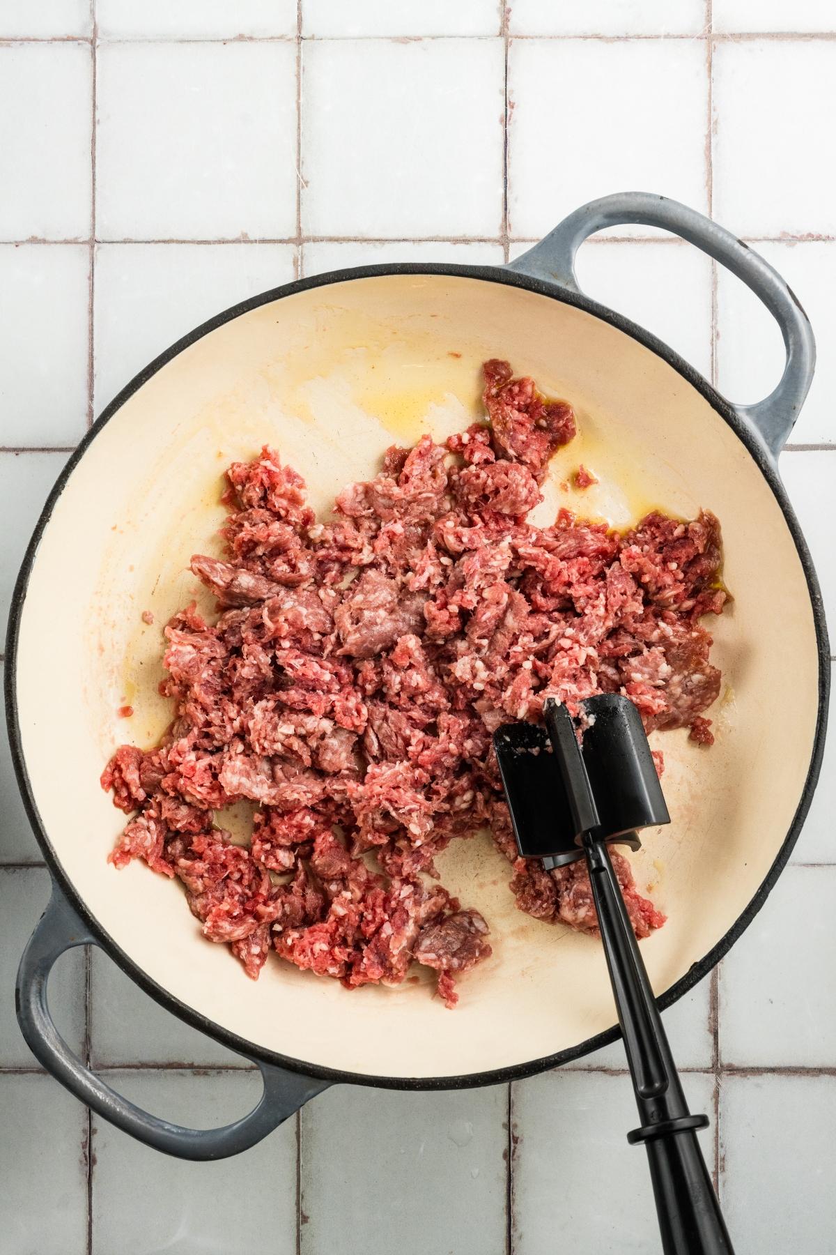 Ground beef being browned in a large pot with a black meat chopper on a white tiled surface.