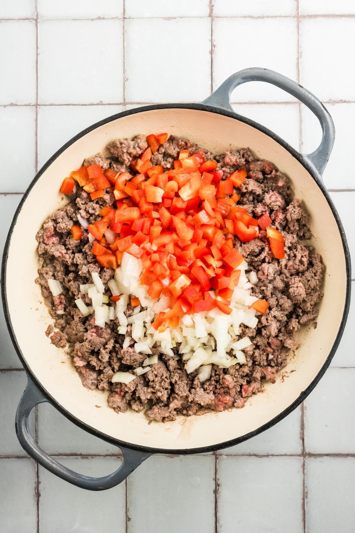 A pot with cooked ground beef, chopped onions, and diced red bell peppers on a white tiled surface.