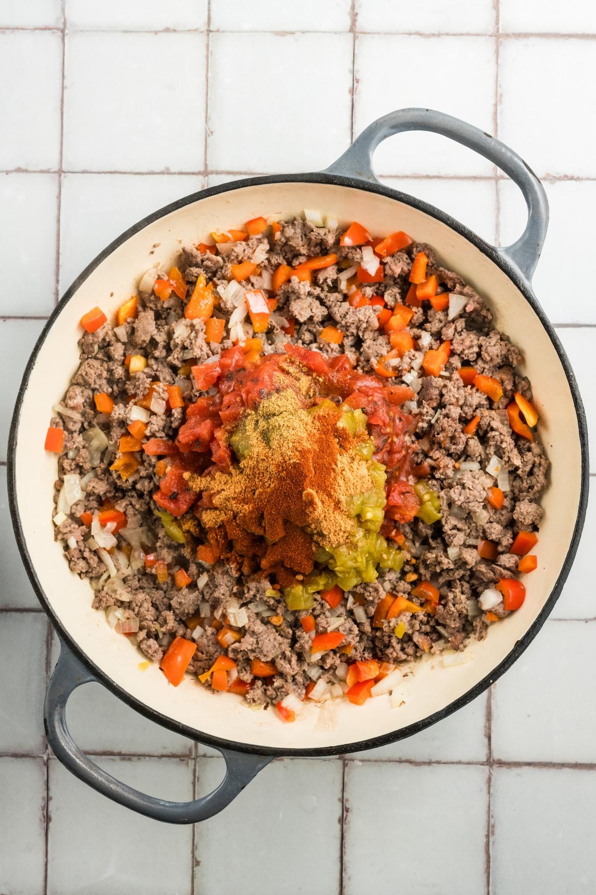 Large pot with ground beef, chopped peppers, onions, tomatoes, and spices ready to be mixed on a tiled surface.
