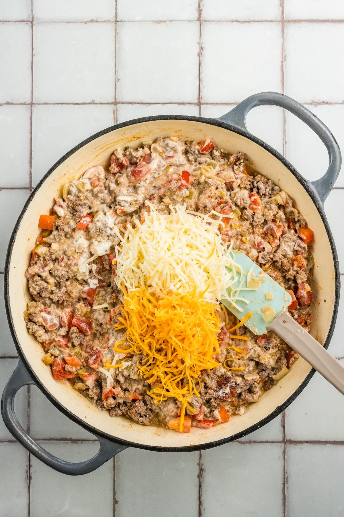 A skillet with ground meat, diced vegetables, and shredded cheese being stirred with a spatula.