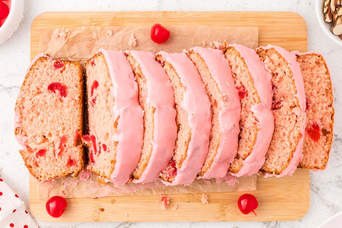 Sliced Cherry Bread with pink icing sits on a wooden board, beautifully garnished with whole cherries.
