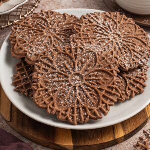 Chocolate pizzelle cookies, dusted with powdered sugar, are arranged on a white plate atop a wooden board for an inviting treat.