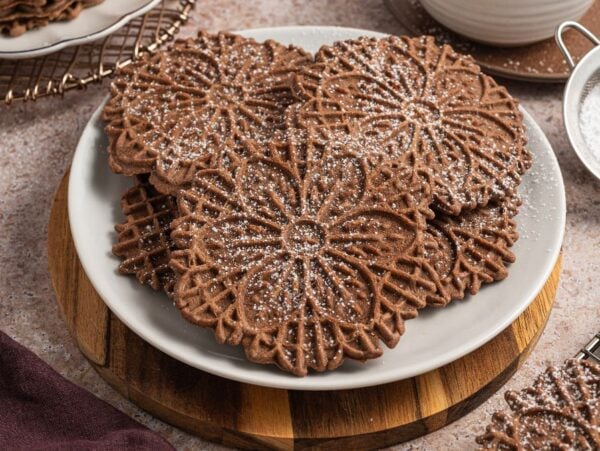 Chocolate pizzelle cookies, dusted with powdered sugar, are arranged on a white plate atop a wooden board for an inviting treat.