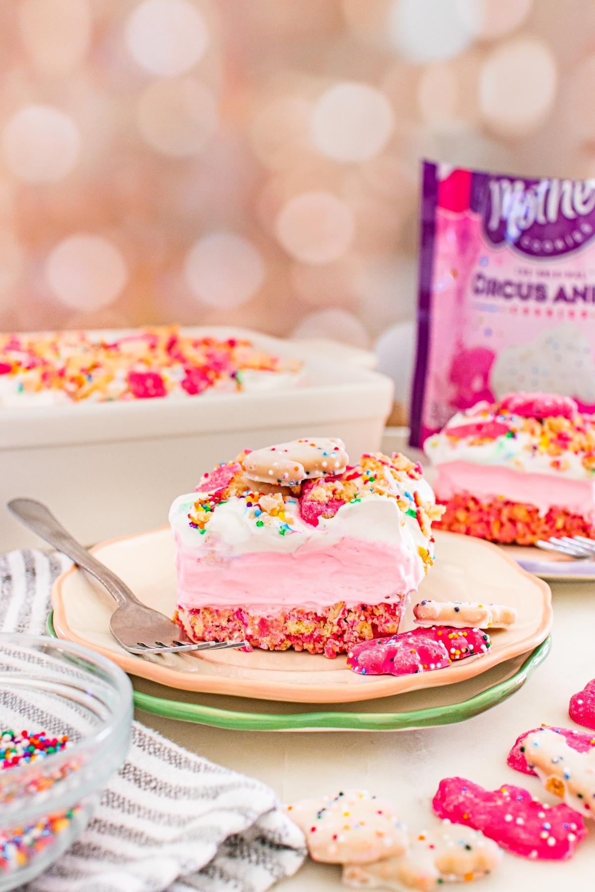 A slice of pink layered dessert topped with frosted animal cookies and sprinkles on a plate.