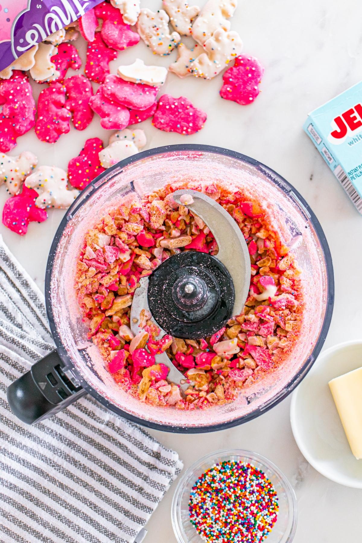 Crushed frosted animal cookies in a food processor with sprinkles, butter, and whole cookies nearby.