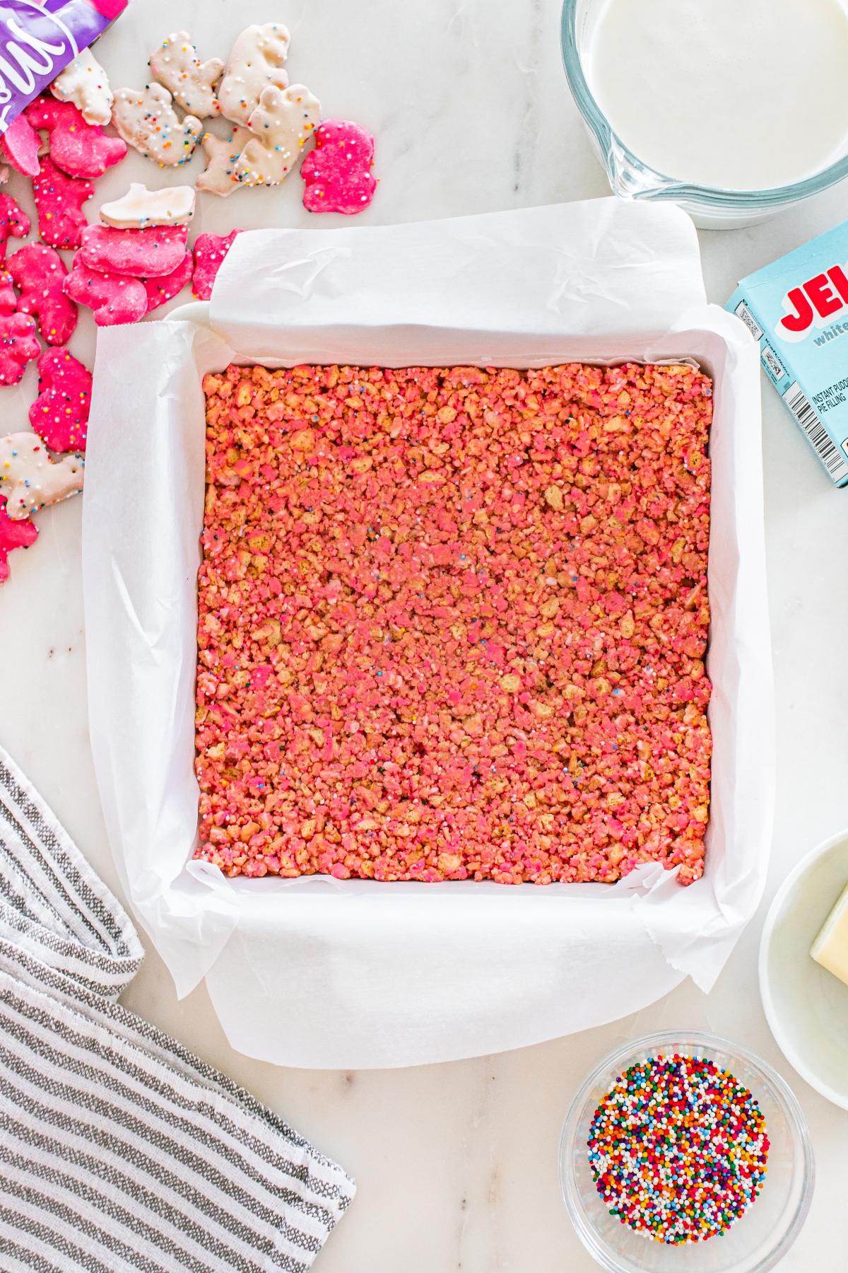 A square pan of pink rice crispy treats on parchment paper, surrounded by sprinkles and frosted animal cookies.