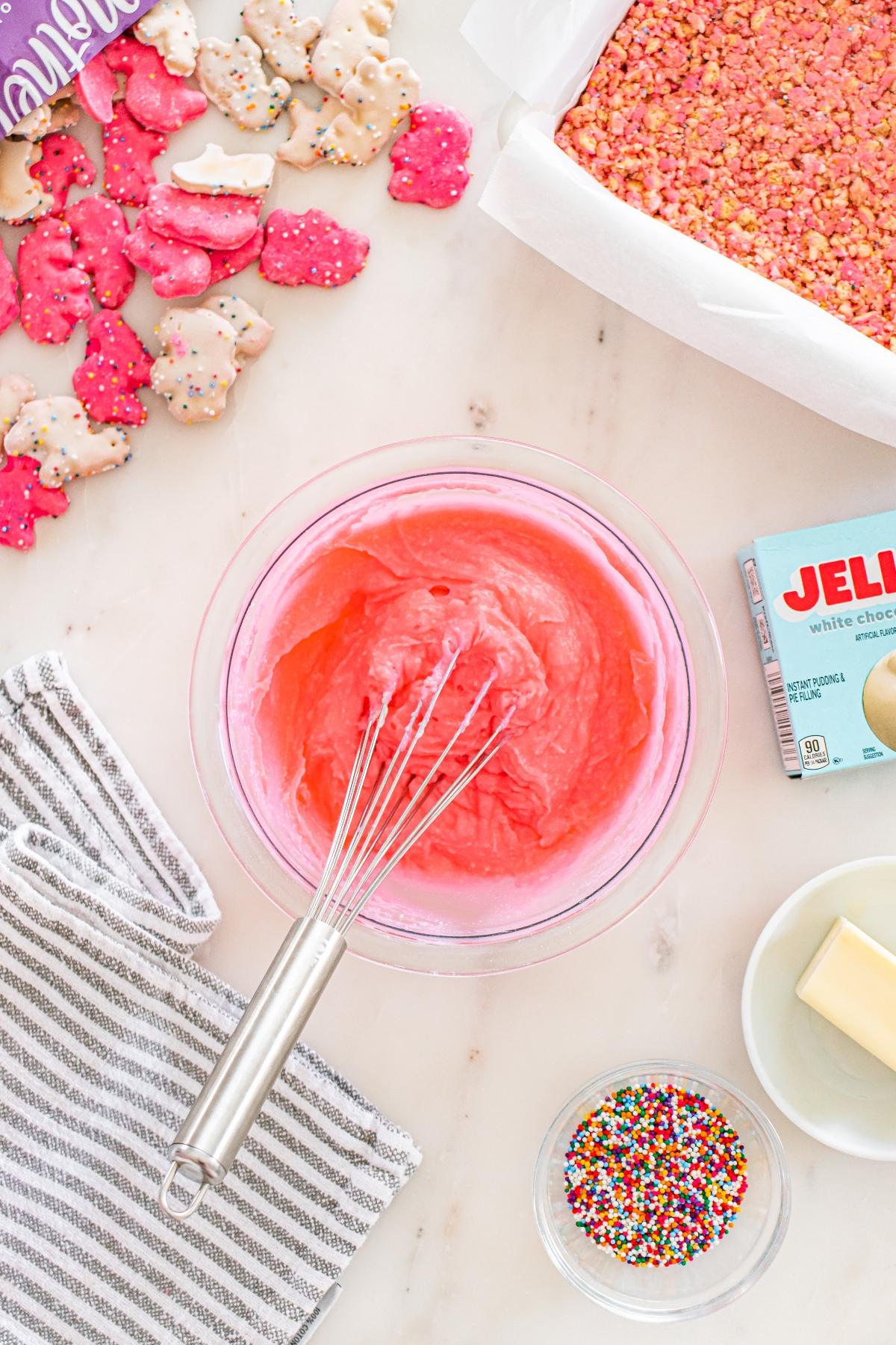A bowl of pink frosting with a whisk, surrounded by sprinkles, cookies, butter, and a striped towel.