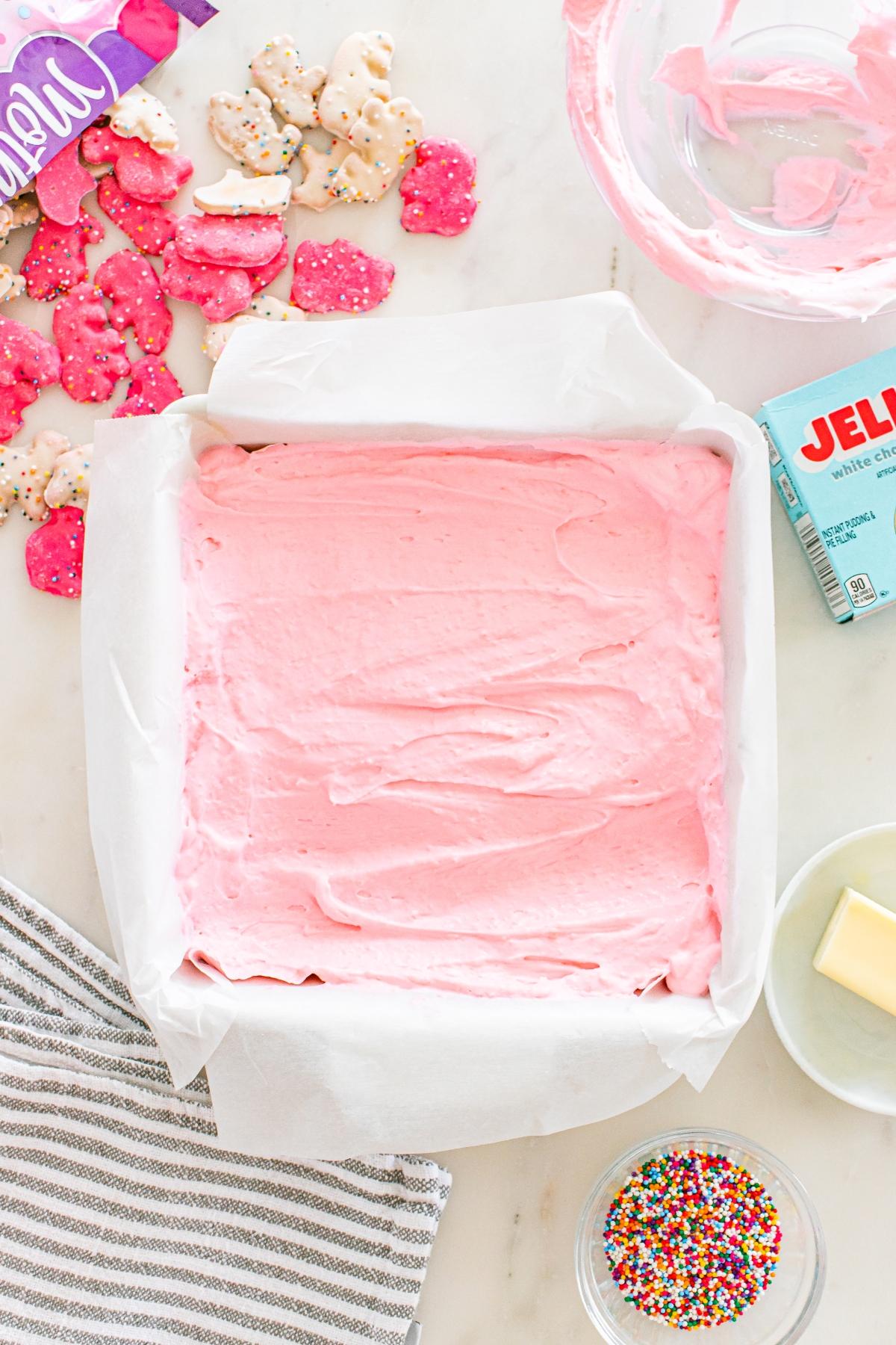 A square pan of pink frosted dessert with sprinkles, animal crackers, and baking ingredients nearby.
