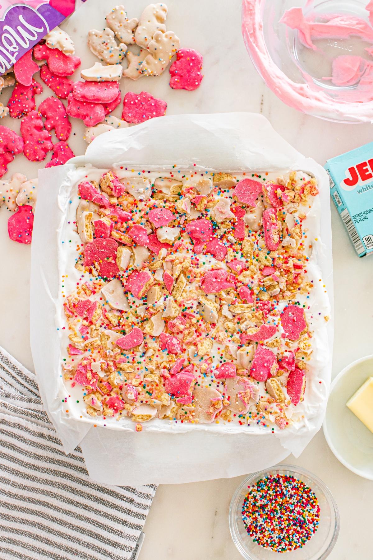 A baking pan of white frosted dessert topped with pink cookies, sprinkles, and animal crackers.