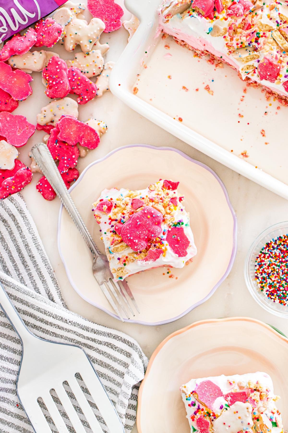 A plate with a dessert topped with frosted animal cookies and sprinkles, next to a fork and baking dish.