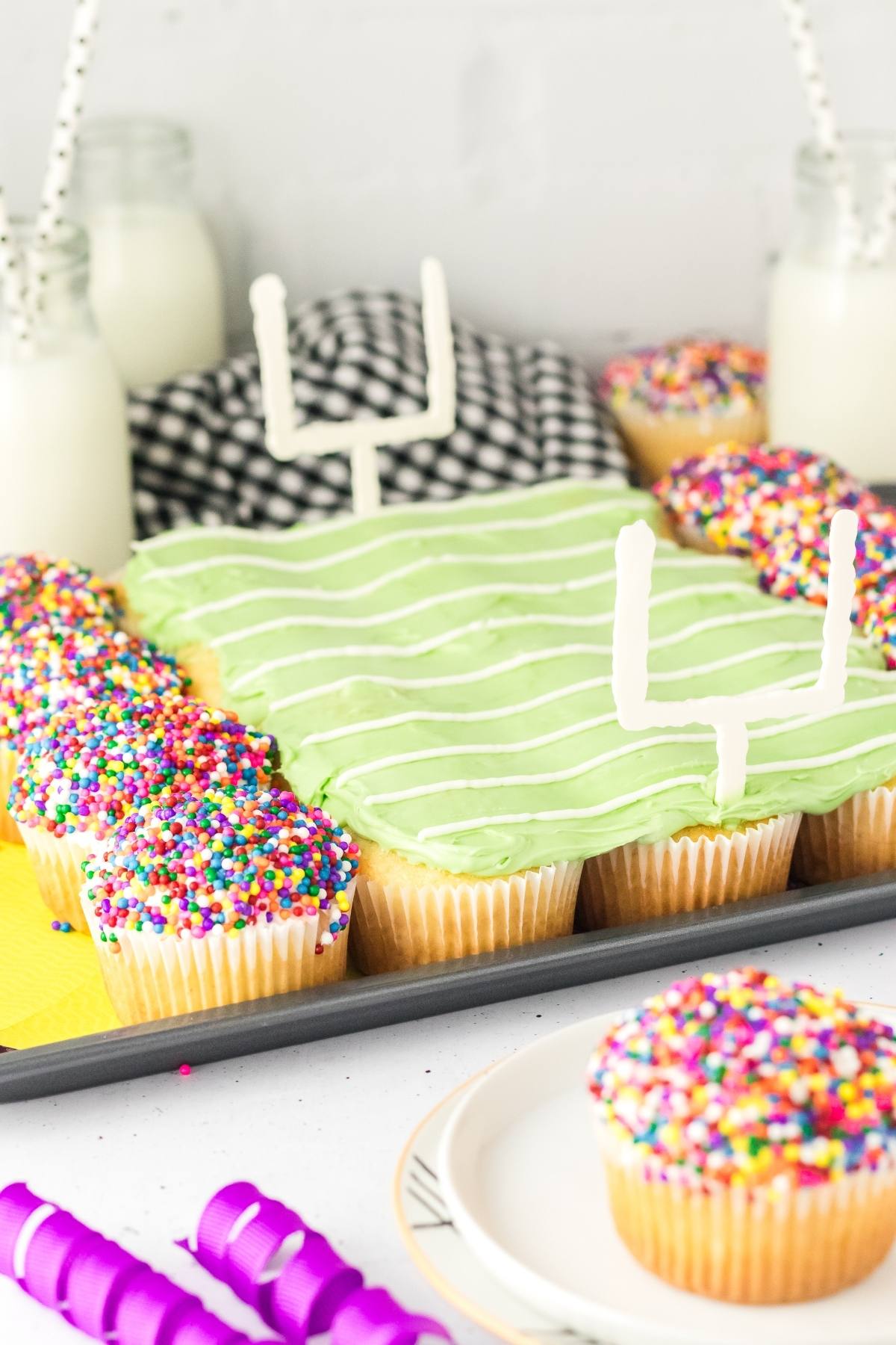 Football stadium cupcakes arranged and decorated as a football field with colorful sprinkles and goal posts, milk in the background.