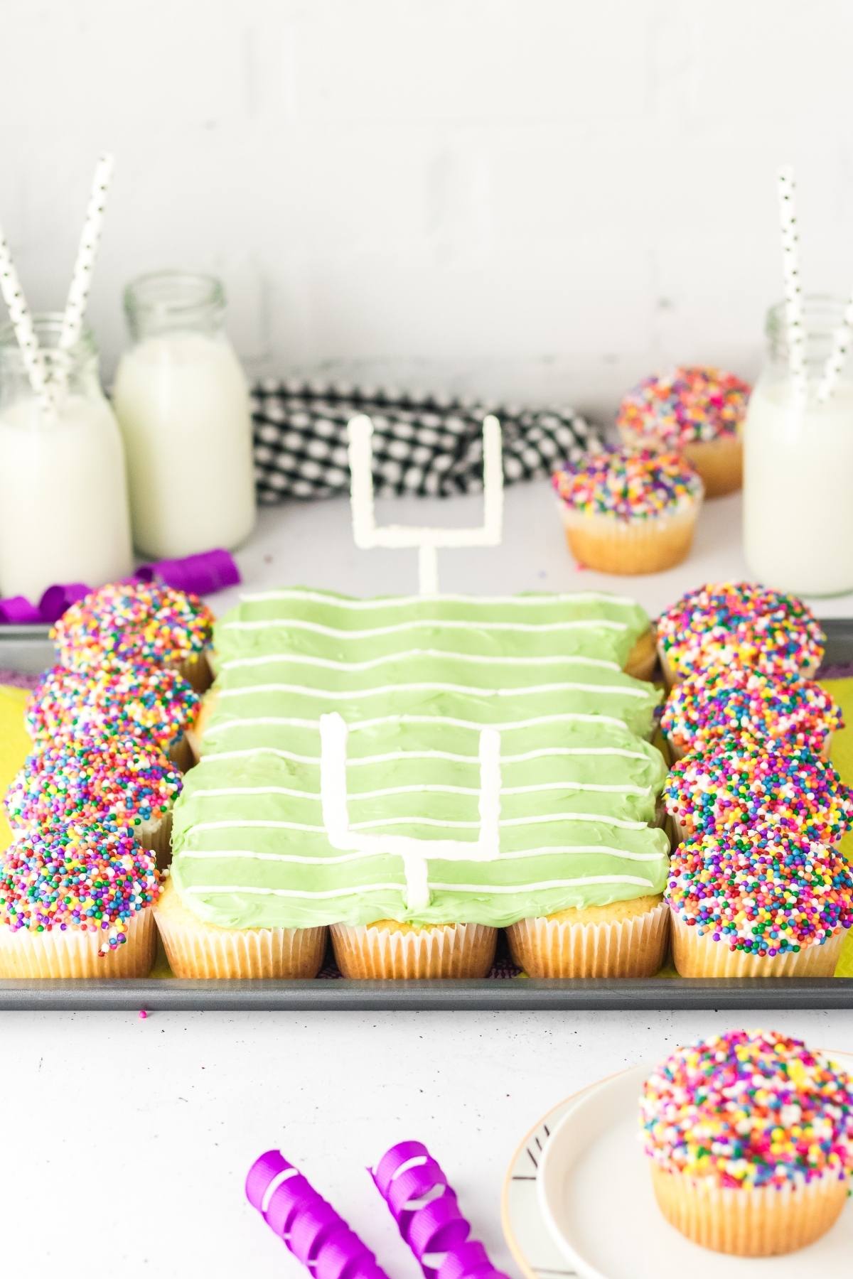 Football stadium cupcakes arranged and decorated like a football field, with colorful sprinkles and bottles of milk in the background.