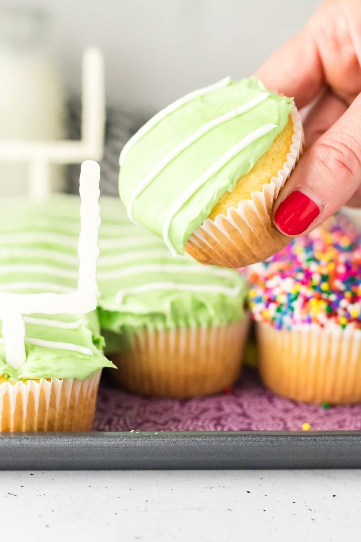 A hand with red nails holds a cupcake with green frosting above a tray of decorated cupcakes.
