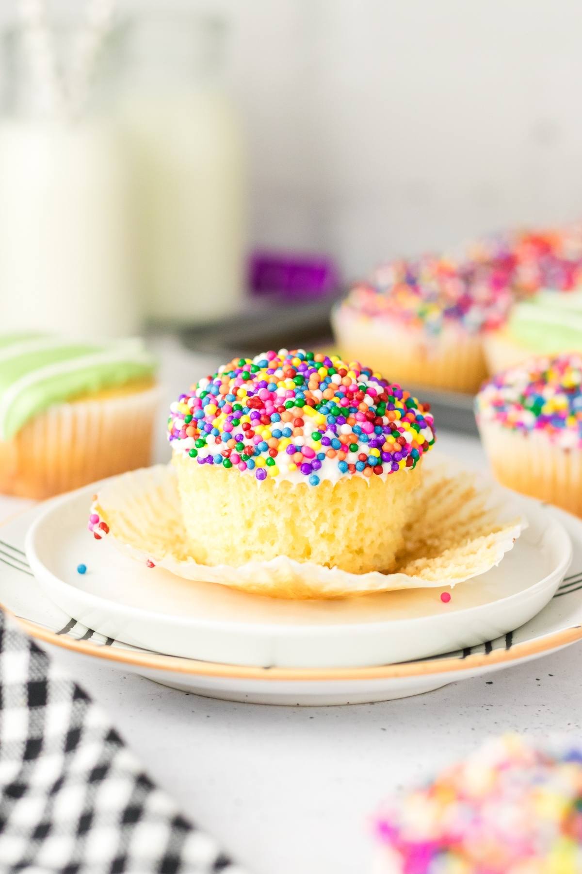A vanilla football stadium cupcake with white frosting and rainbow sprinkles sits on a plate with the wrapper peeled back.