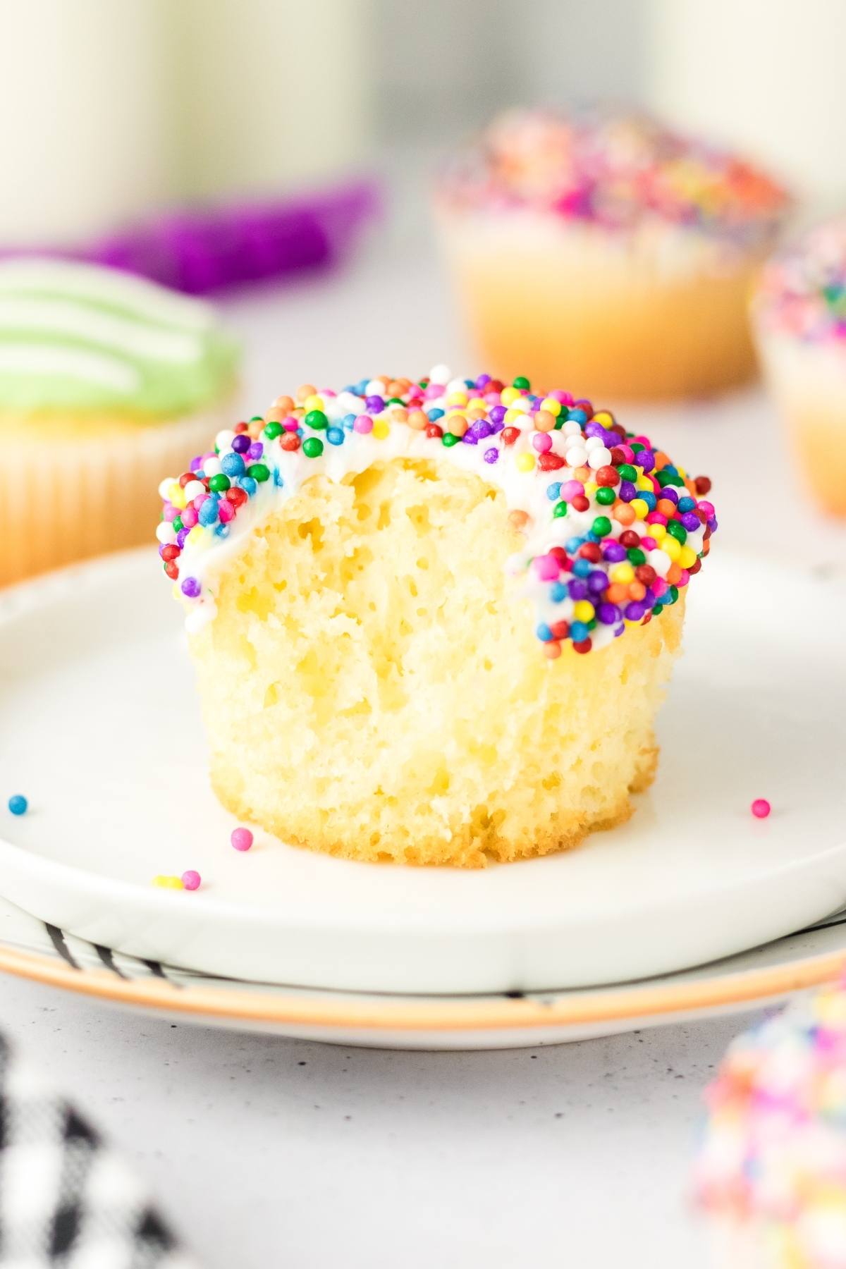 A vanilla football stadium cupcake with white frosting and rainbow sprinkles, missing a bite, sits on a white plate.