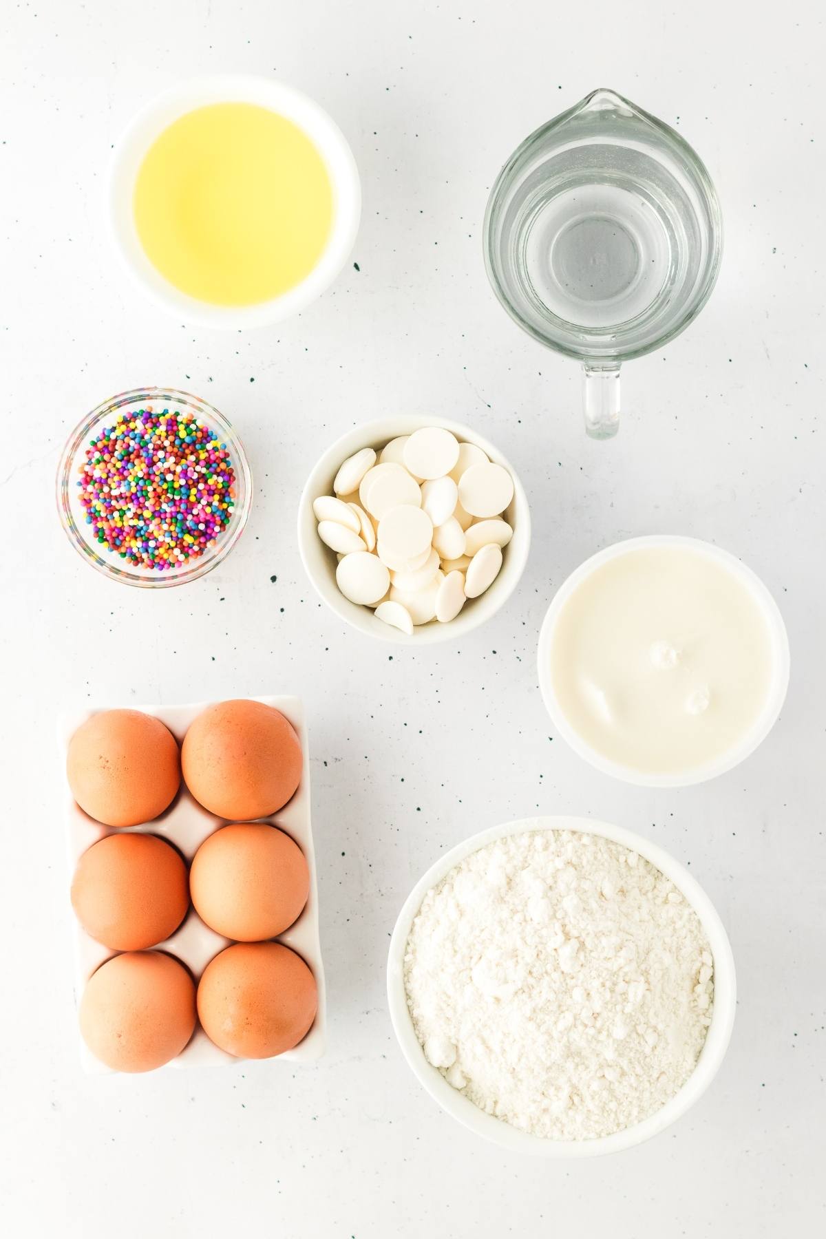 Baking ingredients: eggs, flour, sprinkles, white chocolate, oil, water, and cream on a white background.