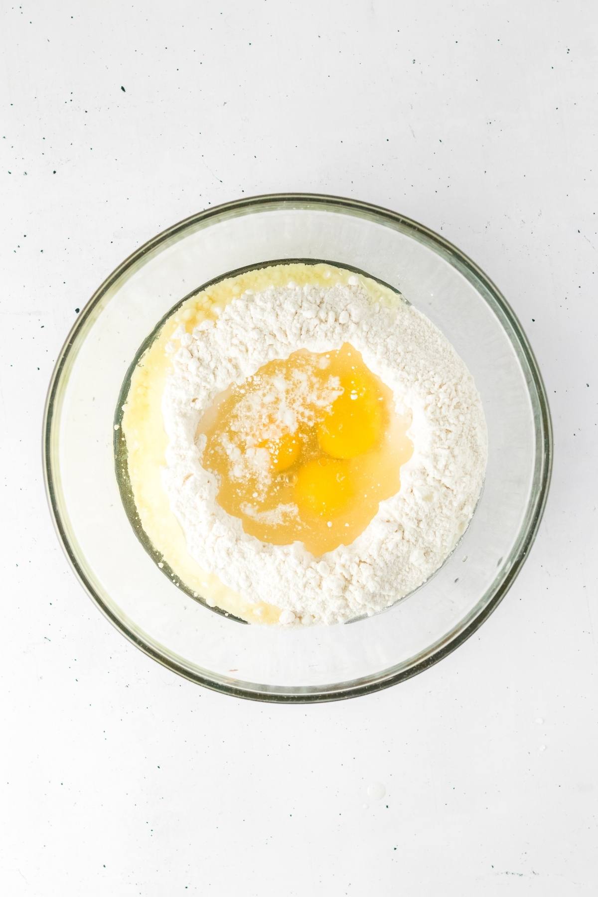 A glass bowl with flour, eggs, and liquid ingredients ready to be mixed on a white surface.
