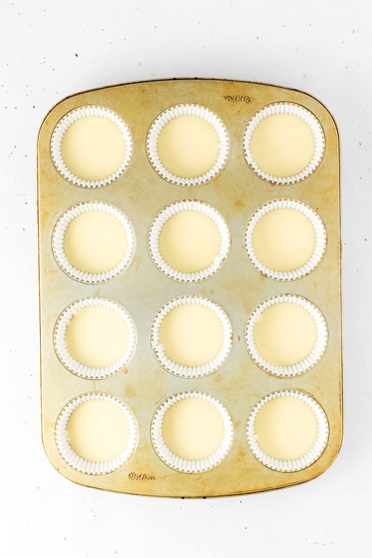 A muffin tin with 12 white cupcake liners filled with pale batter, viewed from above on a white surface.