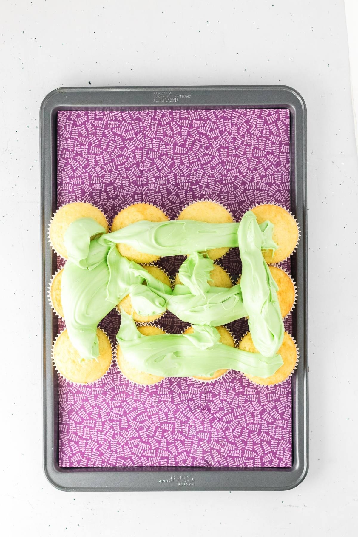 Cupcakes arranged on a tray with green frosting spread on top, sitting on a purple patterned surface.