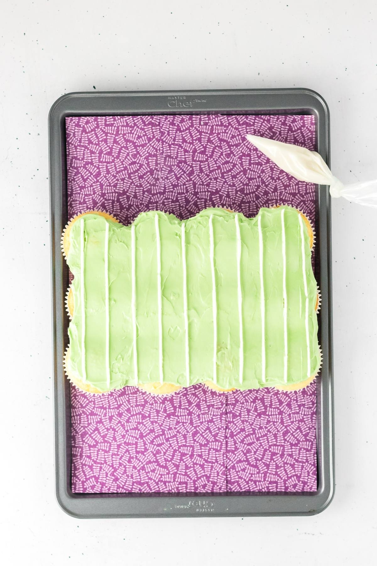 A tray of cupcakes topped with green frosting and white lines, with a piping bag nearby on a purple mat.