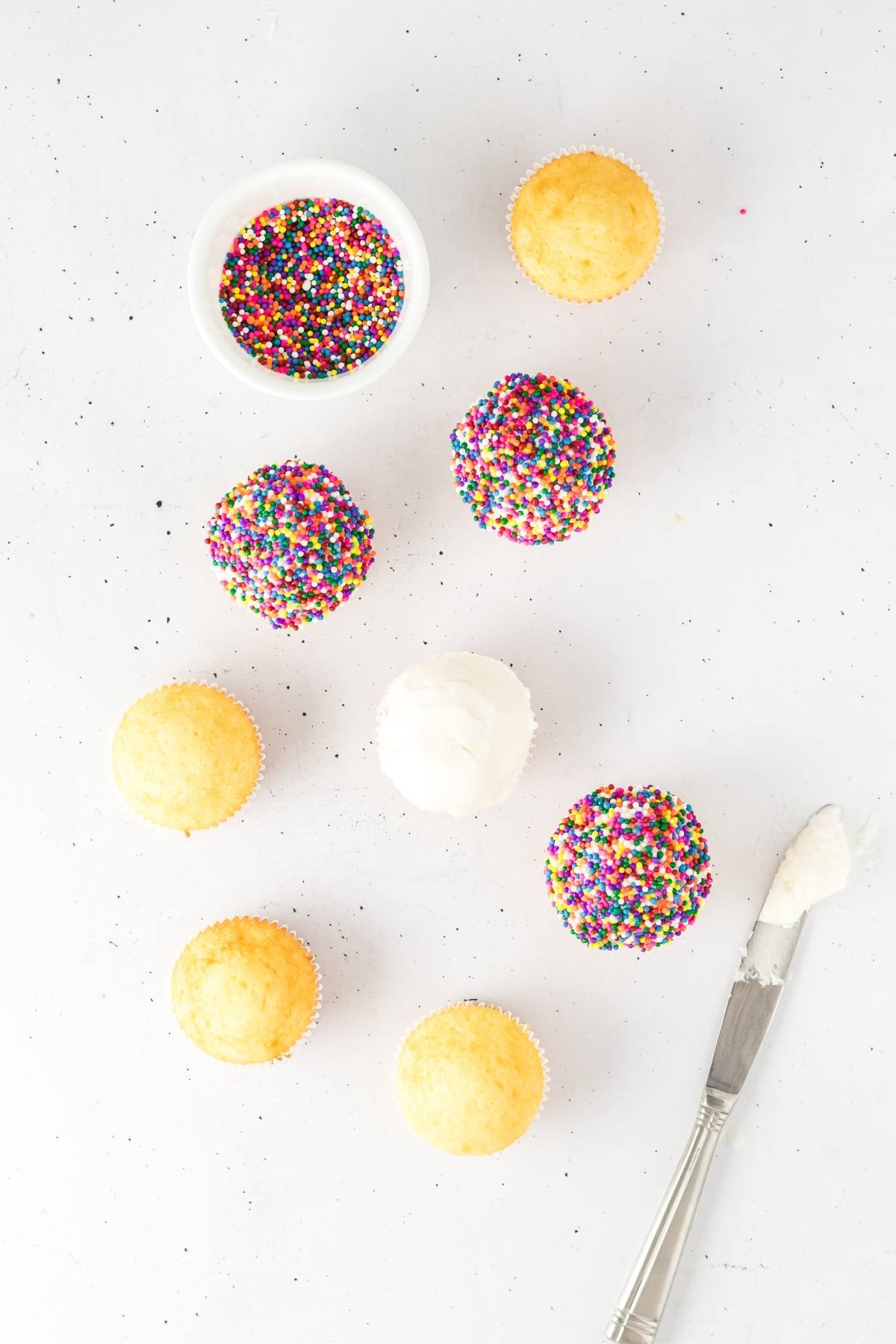 Cupcakes, some plain and some with rainbow sprinkles, a bowl of sprinkles, frosting, and a knife on a white surface.