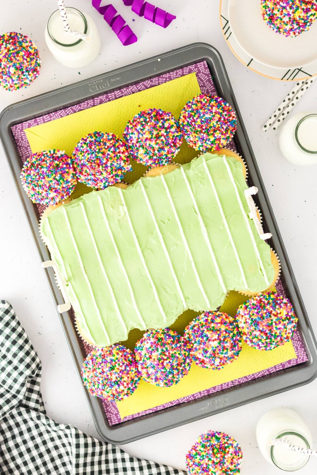 Cupcakes with colorful sprinkles surround a green frosted cupcake cake shaped like a rectangular suitcase.