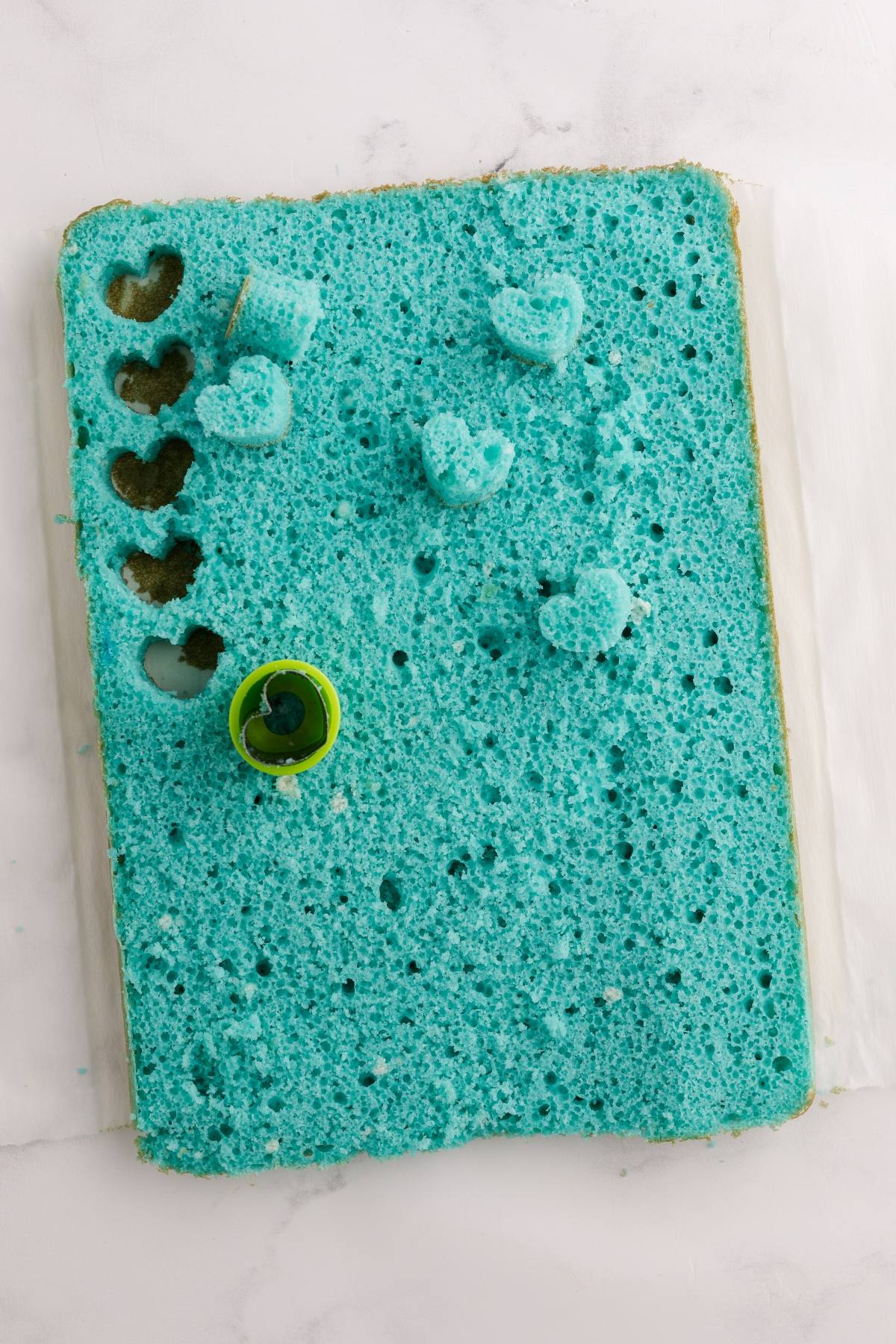 A blue sheet cake with heart shapes cut out and a small heart-shaped cutter on top.