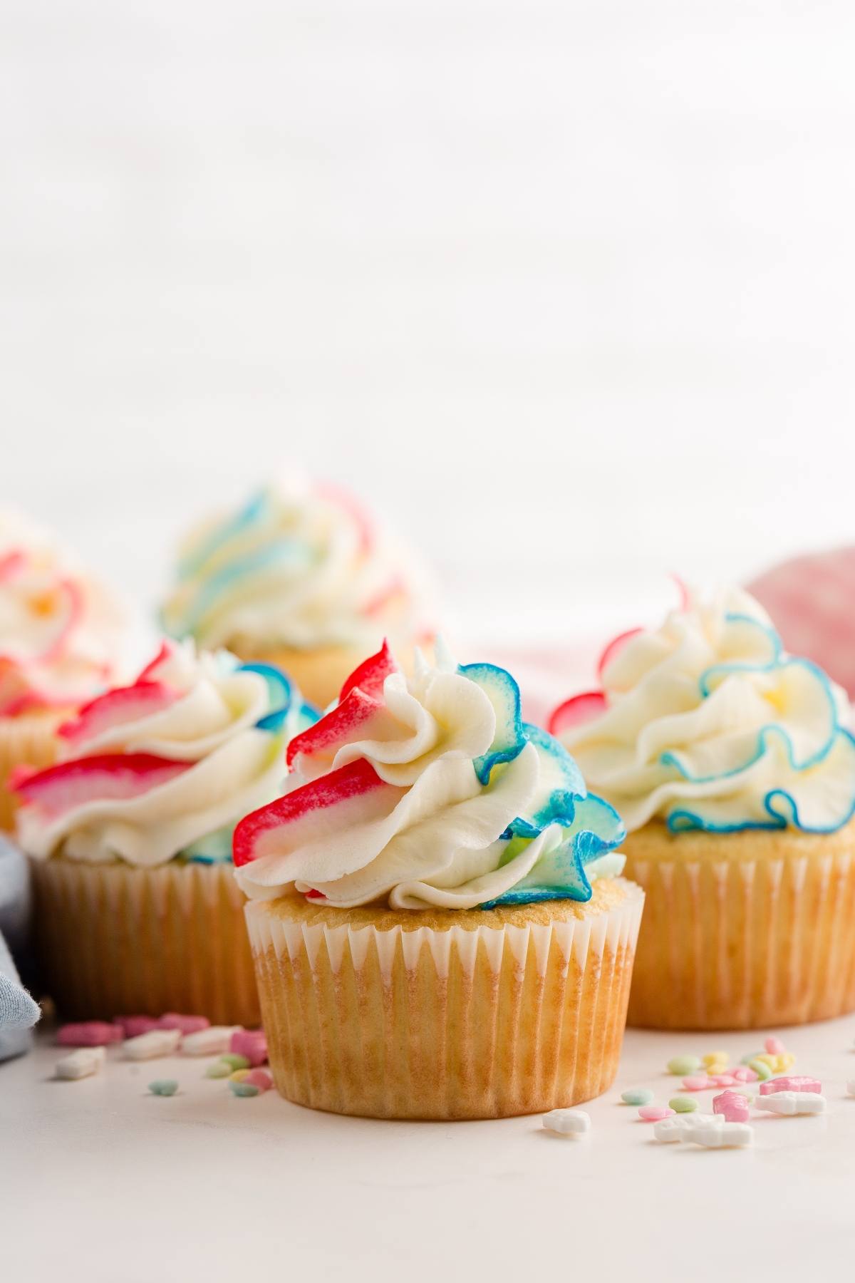 Vanilla gender reveal  cupcakes with white frosting and red and blue swirled edges, with pastel sprinkles nearby.