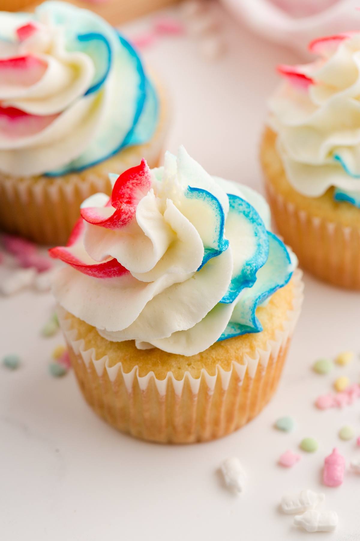 Vanilla Gender reveal  cupcakes topped with swirled red, white, and blue frosting, surrounded by pastel sprinkles.