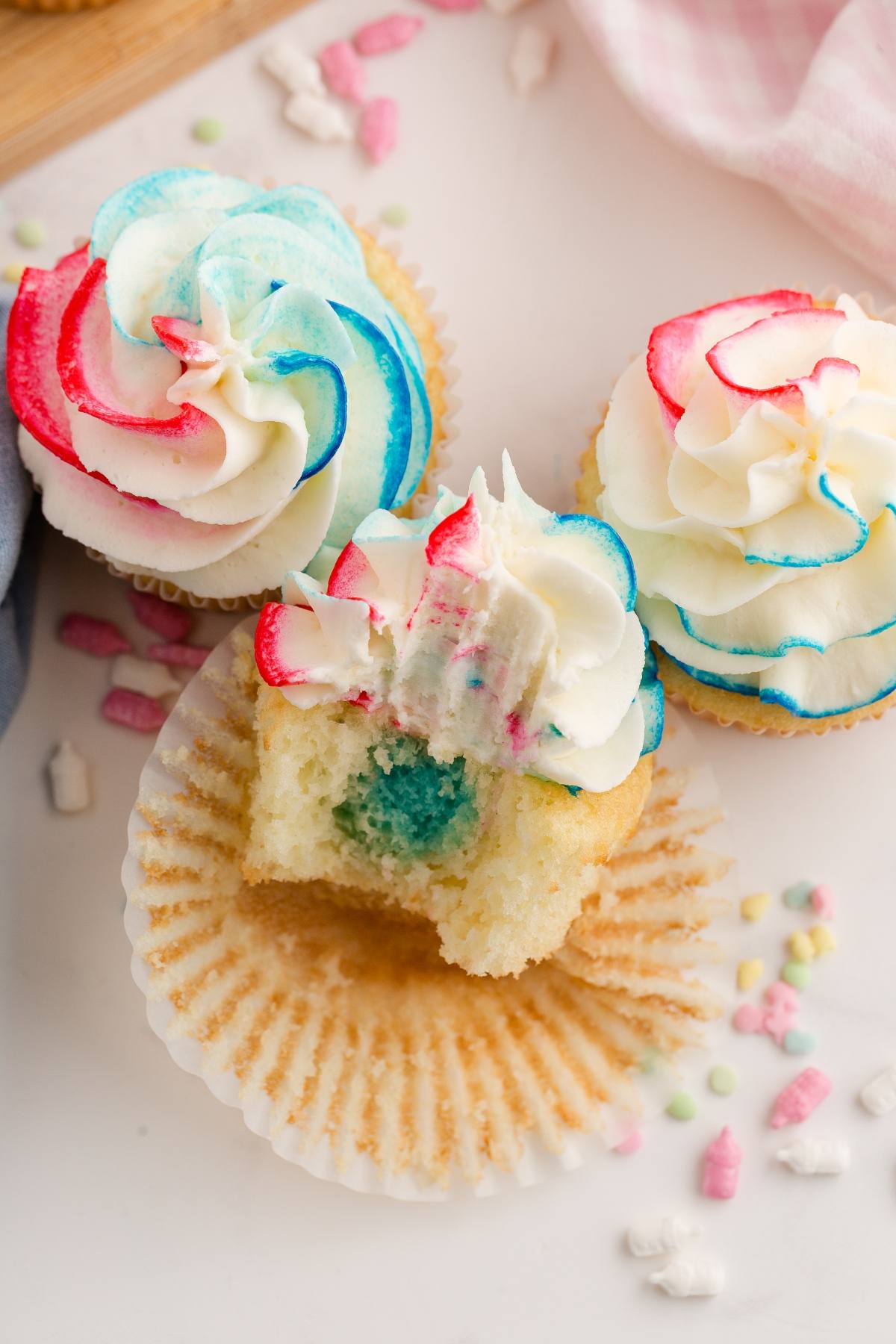 Gender reveal cupcakes with red, blue, and white swirled frosting; one has a bite revealing blue filling inside.